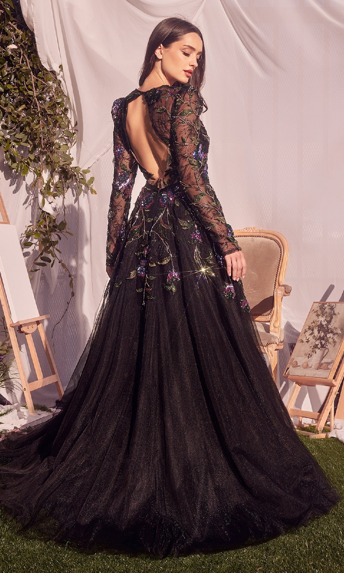 A woman in the Andrea & Leo MA107 Long-Sleeve Black Formal Dress stands in a decorated room with chairs, plants, and draped white fabric.